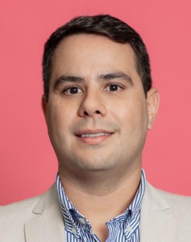 A man with short dark hair is wearing a light beige blazer and a blue and white striped shirt against a pink background.